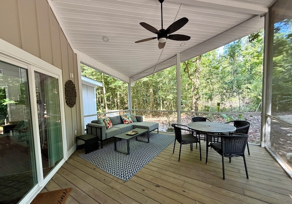 A covered outdoor area with a ceiling fan, a sofa set, a dining table with chairs, and a view of a forested area.