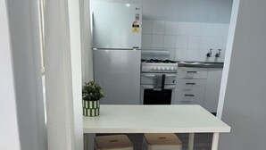 Een compacte keuken met een wit aanrecht, een gasfornuis en een koelkast. Er staan vier houten krukken onder het aanrecht.