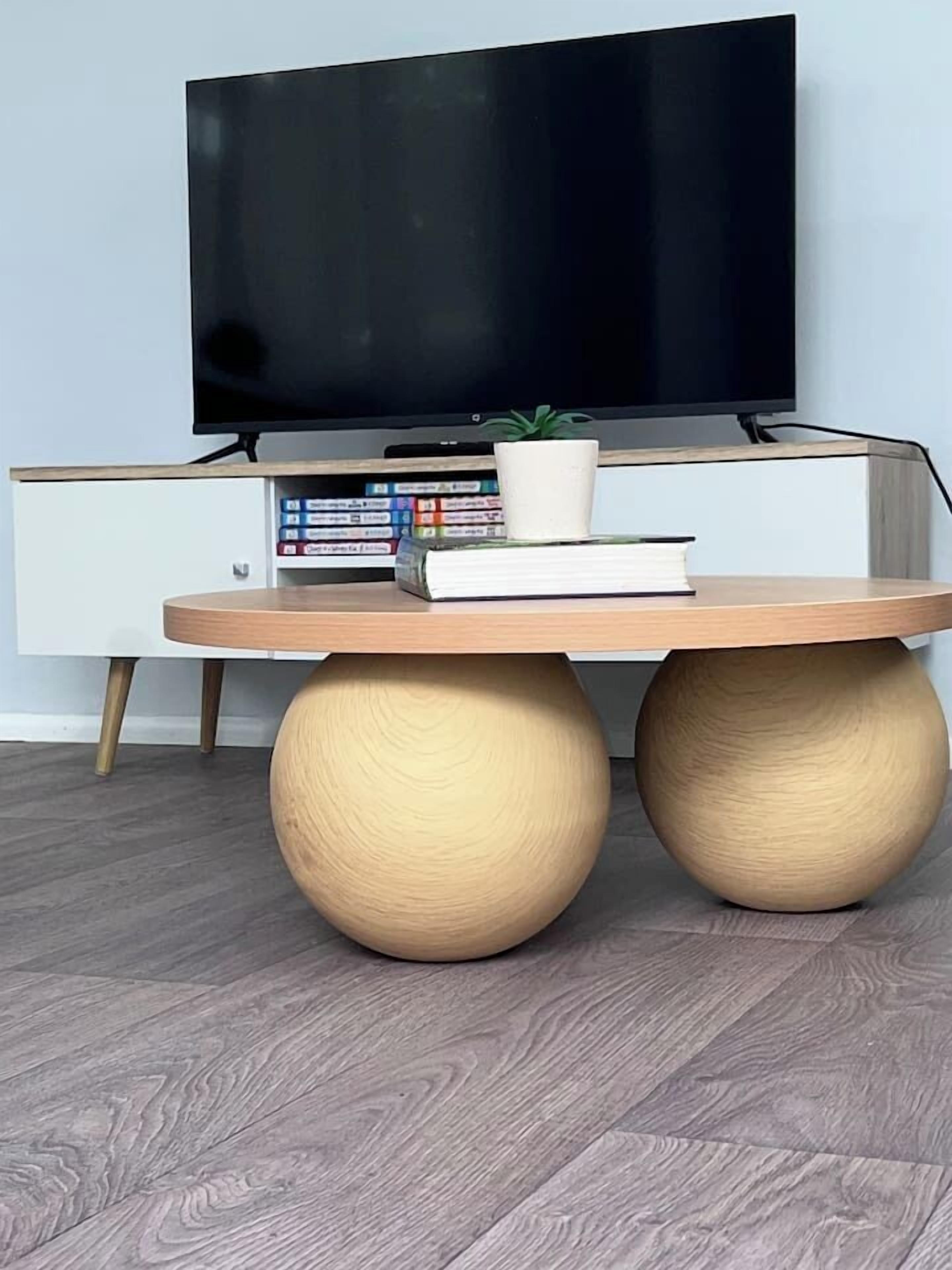 A modern room with a flat-screen TV mounted on a white cabinet, a round wooden table with two large legs, and a bookshelf with books and decorative items.