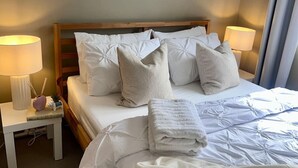 En pænt redt seng med hvide sengetøj og puder, et natbord med en lampe og en lille pynteting.