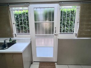 Una cocina con una puerta blanca, dos ventanas con barrotes, un fregadero y un ventanal pequeño debajo de la puerta.