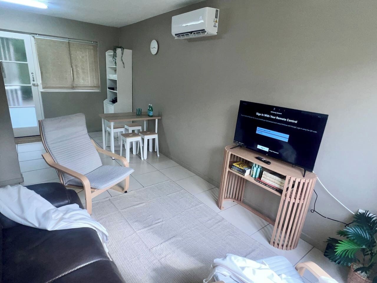 Ein Wohnzimmer mit einer Couch, einem Fernseher auf einem Holzboden, einem kleinen Tisch mit Stühlen und einer Klimaanlage an der Wand.