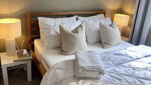 A neatly made bed with white linens and pillows, a bedside table with a lamp, and a small decorative item. - u4-Traditional Unit-Quiet Close CBD-AirConSmartTV (Norman Park)