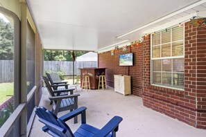 A covered outdoor seating area with chairs and a table, a brick wall with a sign, and a window with blinds. - Coastal Orange Beach Retreat | Minutes to the Beach, The Wharf & Family Fun (Orange Beach)