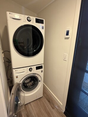 A stacked washer and dryer unit in a corner of a room with a wooden floor and a door on the right.