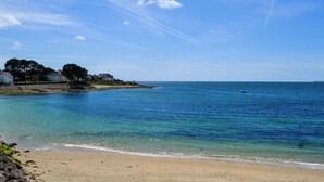 En strand med klart vatten, en sandstrand och en rad hus i bakgrunden.