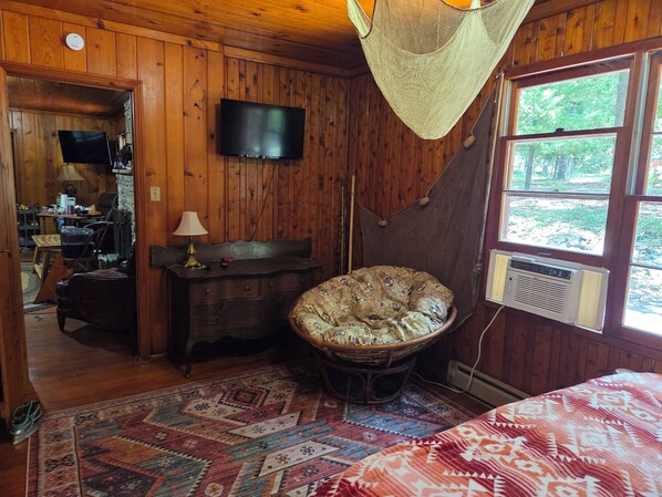 A wooden cabin interior with a bed, a television, a hammock, and a large window. - NEW! 3Br Retro-style Cabin on KY Lake waterfront (Gilbertsville)