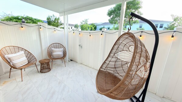 Un patio cubierto con sillas de mimbre y una hamaca colgante, suelo de baldosas blancas y una valla blanca con luces de cuerda.