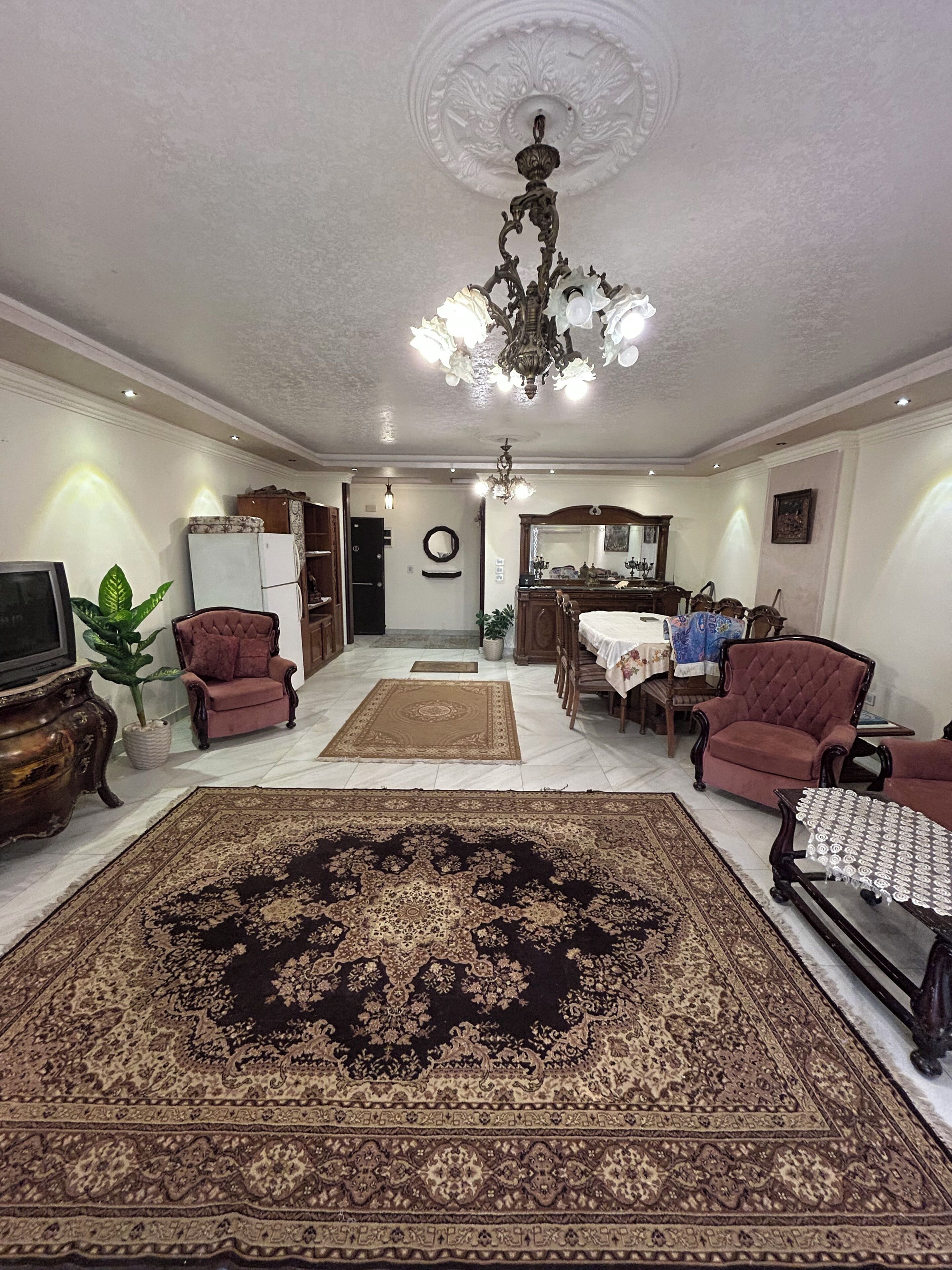 A spacious living room with a large ornate rug, a chandelier, and a dining area with a table and chairs.