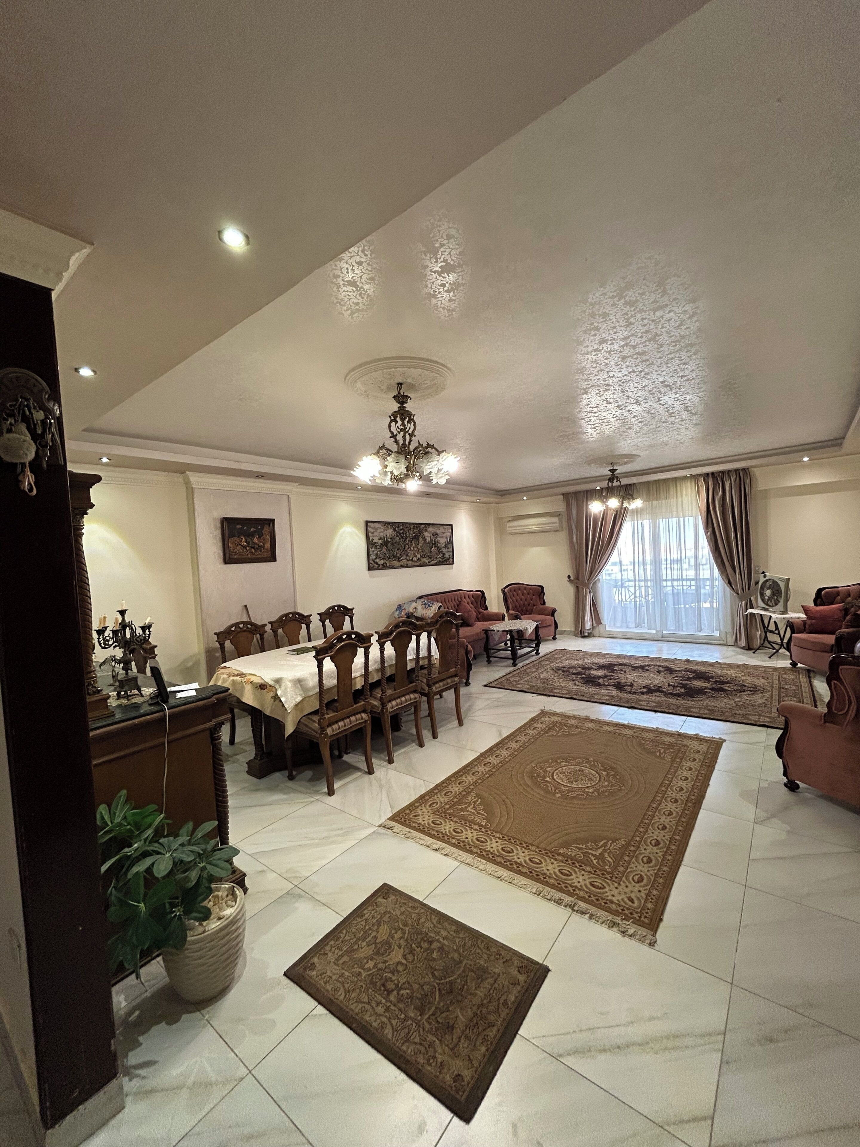 A spacious living room with a dining area, a chandelier, and a large window with curtains.