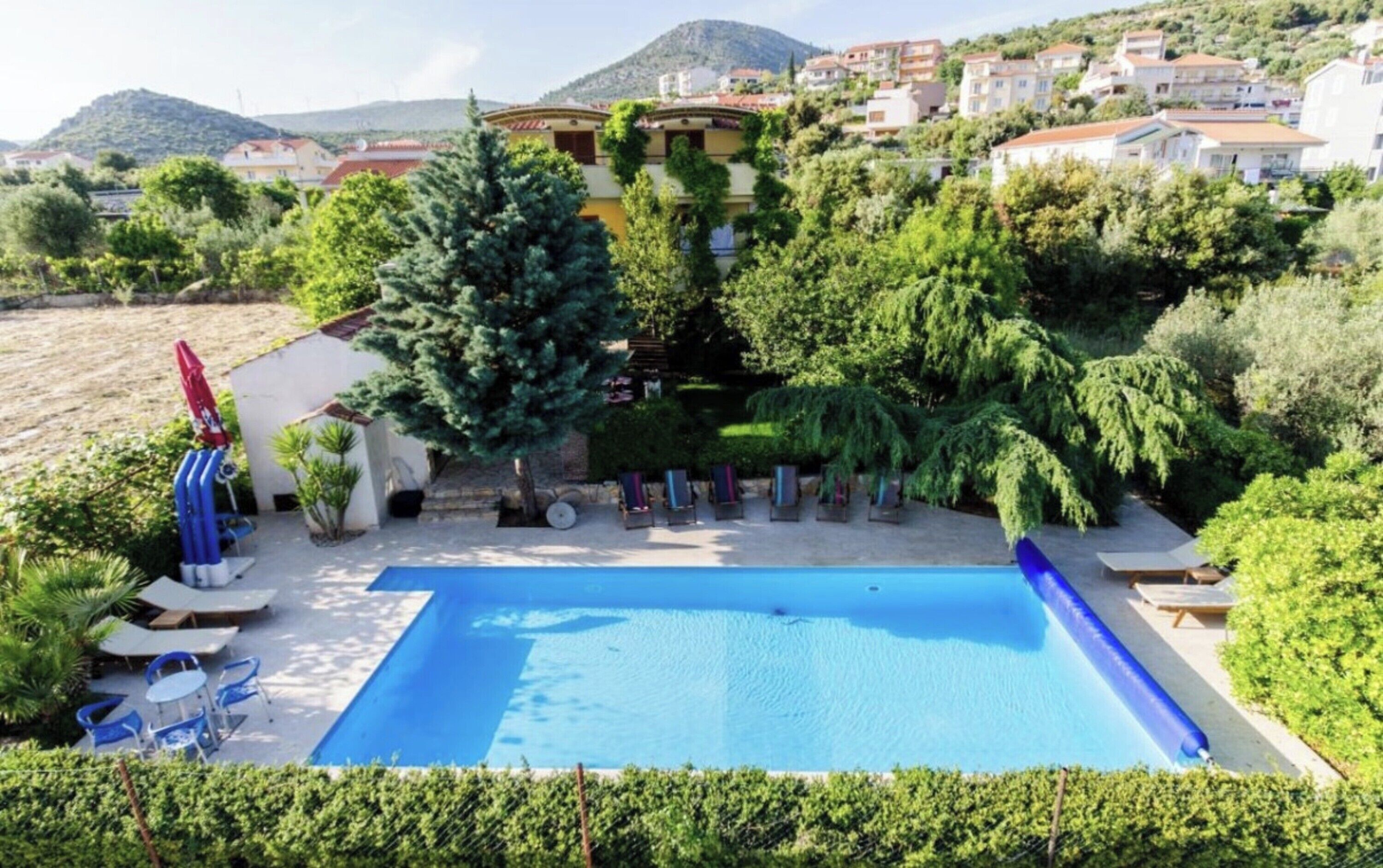 A swimming pool with lounge chairs and umbrellas, surrounded by greenery and buildings in the background.