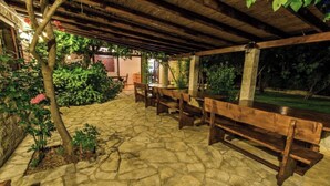 An outdoor patio area with stone flooring, wooden benches, and a covered section with a thatched roof. - A 1  apt w shared pool and balcony, near the beach (Seget Vranjica)