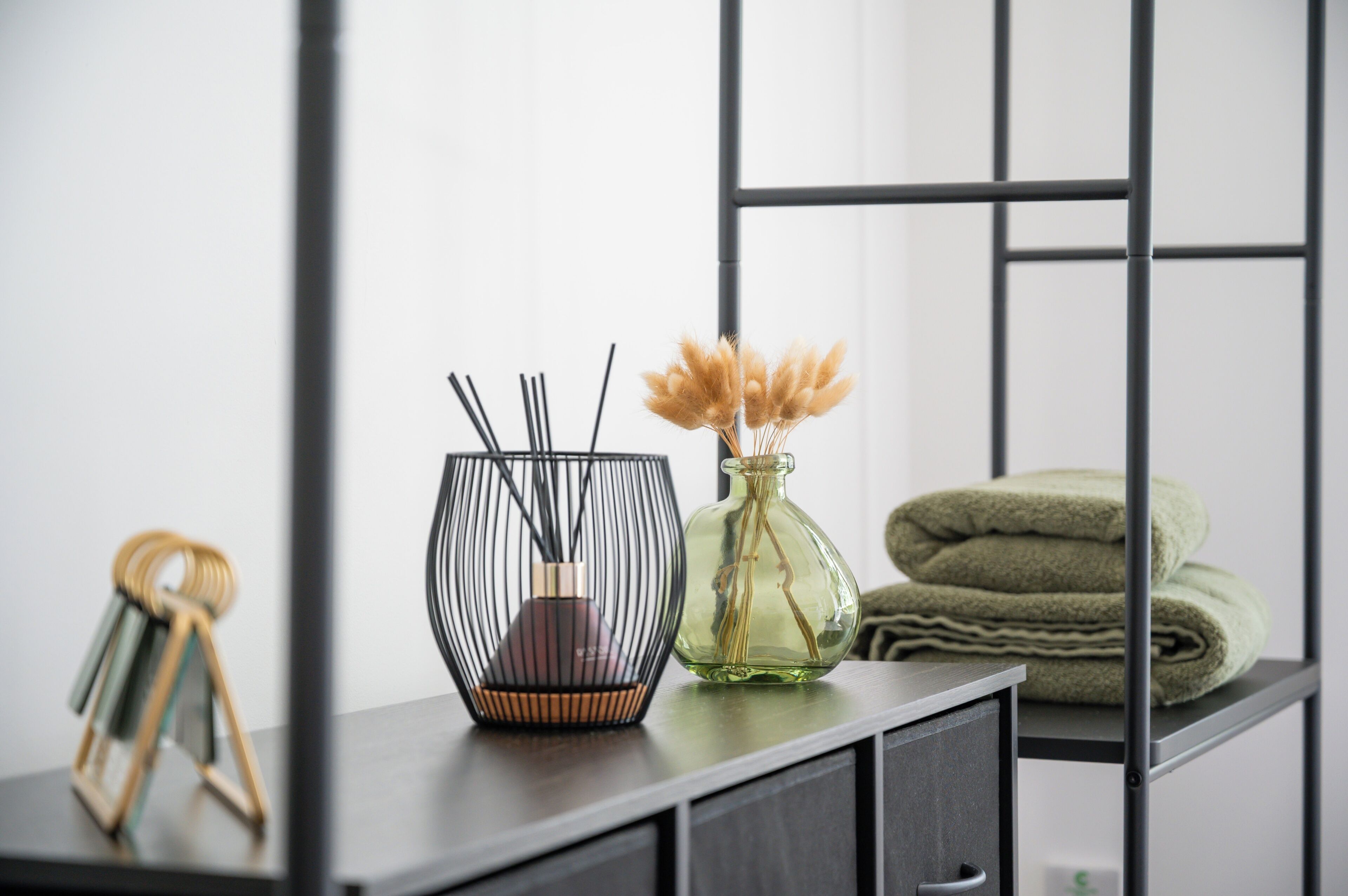 A modern, minimalist room with a dark wooden desk, a black wireframe vase with dried pampas grass, a clear glass vase with pampas grass, and neatly folded green towels.