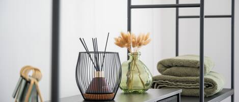 A modern, minimalist room with a dark wooden desk, a black wireframe vase with dried pampas grass, a clear glass vase with pampas grass, and neatly folded green towels.
