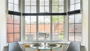A room with a table set for two, two chairs, a window with blinds, and a view of a brick wall and greenery outside. - Emerald Garden Suite - Wifi,Parking,Garden,Sleep 2 (London)