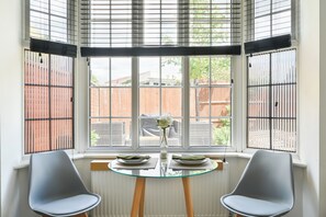 Quarto com mesa posta para dois, duas cadeiras, janela com persianas e vista para uma parede de tijolos e vegetação lá fora.