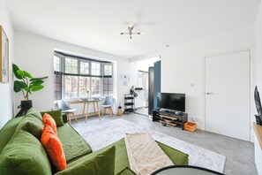 A modern living room with a green sofa, a television, and a large window. - Emerald Garden Suite - Wifi,Parking,Garden,Sleep 2 (London)