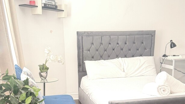 A bedroom with a grey upholstered bed, a blue bench, a potted plant, and a small table with a lamp. - Cozy London Apartment with City Views (London)