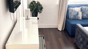 A modern living room with a white desk, a mounted flat-screen TV, a potted plant, a blue sofa with cushions, and a large window with curtains. - Cozy London Apartment with City Views (London)