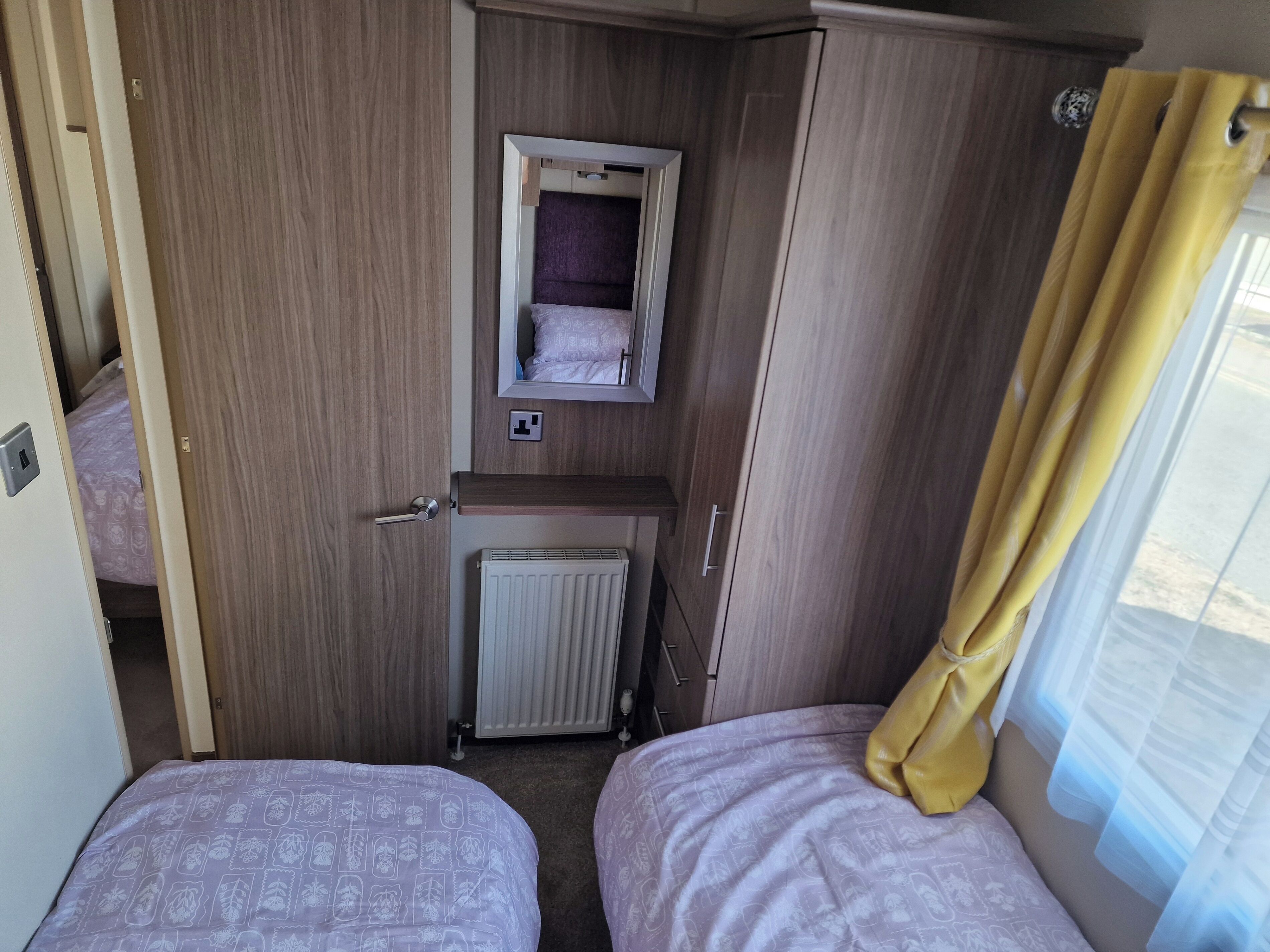Een compacte kamer met twee bedden, een klein raam met gordijnen, een radiator en een spiegel boven een plank.