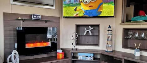 A modern living room with a flat-screen TV mounted on the wall, a fireplace, and a small shelf with decorative items.
