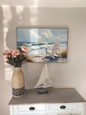 A painting of a beach scene with a sailboat and chair, a woven vase with flowers, and a small sailboat model on a dresser.