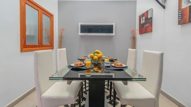 A dining area with a glass table, white chairs, a fruit bowl, and a television on the wall. - Casa Encanto Arinaga (Aguimes)