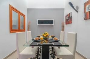 A dining area with a glass table, white chairs, a fruit bowl, and a television on the wall. - Casa Encanto Arinaga (Aguimes)