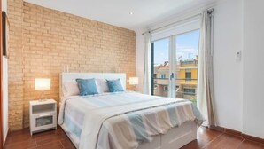 A bedroom with a brick accent wall, a large bed with a striped bedspread, a nightstand with a lamp, and a view of buildings through a large window. - Casa Encanto Arinaga (Aguimes)