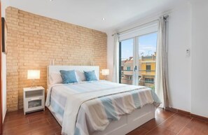 Quarto com parede de tijolos decorativa, uma cama grande com colcha listrada, mesa de cabeceira com abajur e vista para prédios através de uma janela ampla.