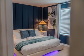 Une chambre à coucher avec un lit, une table de chevet, une lampe et une œuvre d’art accrochée au mur.