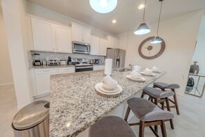 A modern kitchen with white cabinetry, granite countertops, and stainless steel appliances. There are bar stools and a large mirror on the countertop. - Go!#41 - WINDSOR AT WESTSIDE (Kissimmee)