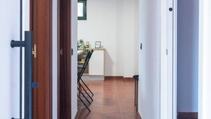 A hallway with a wooden door on the left, a tiled floor, and a view into a room with a window and a small table. - Casa De Invitados El Rincon De Itara (Firgas)