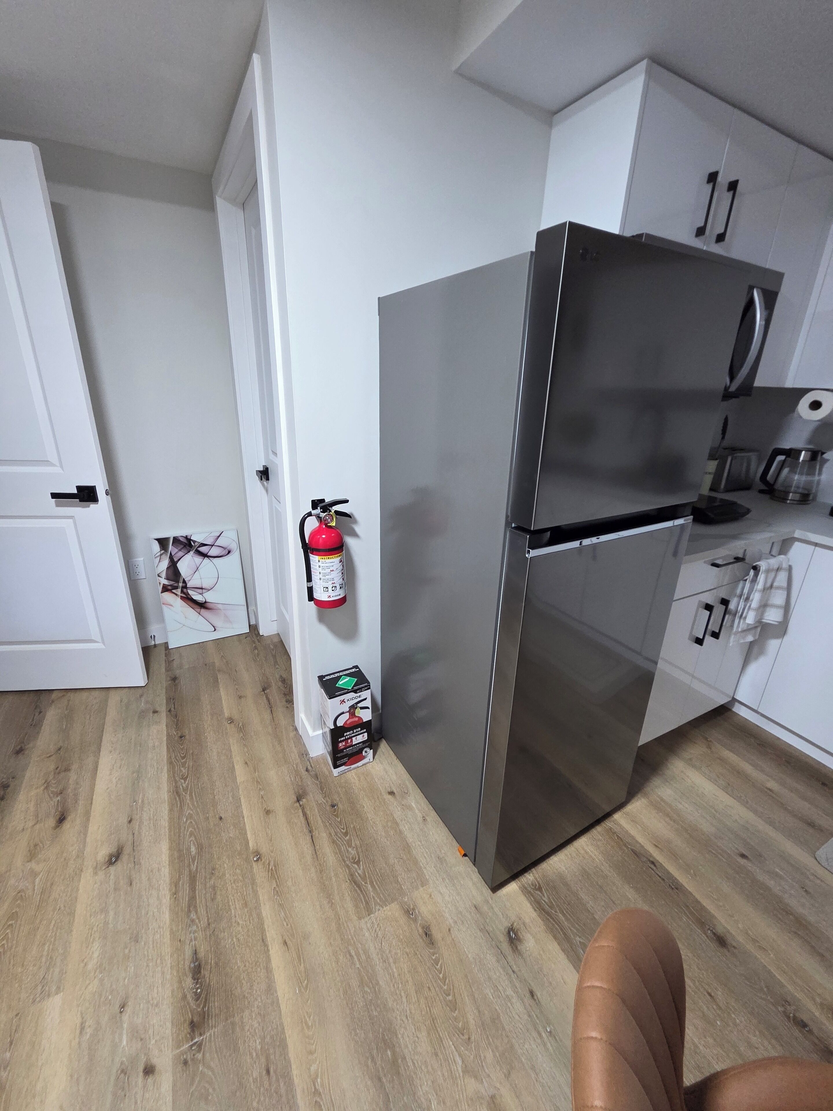 Une cuisine équipée d’un réfrigérateur en acier inoxydable, d’un plancher en bois, de meubles blancs et d’un extincteur fixé au mur.