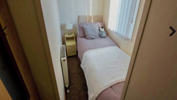 Una habitación de hotel pequeña y estrecha con una cama individual, una mesita de noche, una lámpara y una ventana con cortinas.