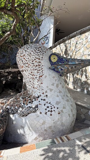 Een mozaïeksculptuur met een vogelmotief, omringd door begroeiing en een betegelde vloer.