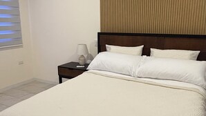 A neatly made bed with a white duvet and pillows, a bedside table with a lamp, and a window with blinds. - Katarma Deluxe Apartments (Puerto Baquerizo Moreno)