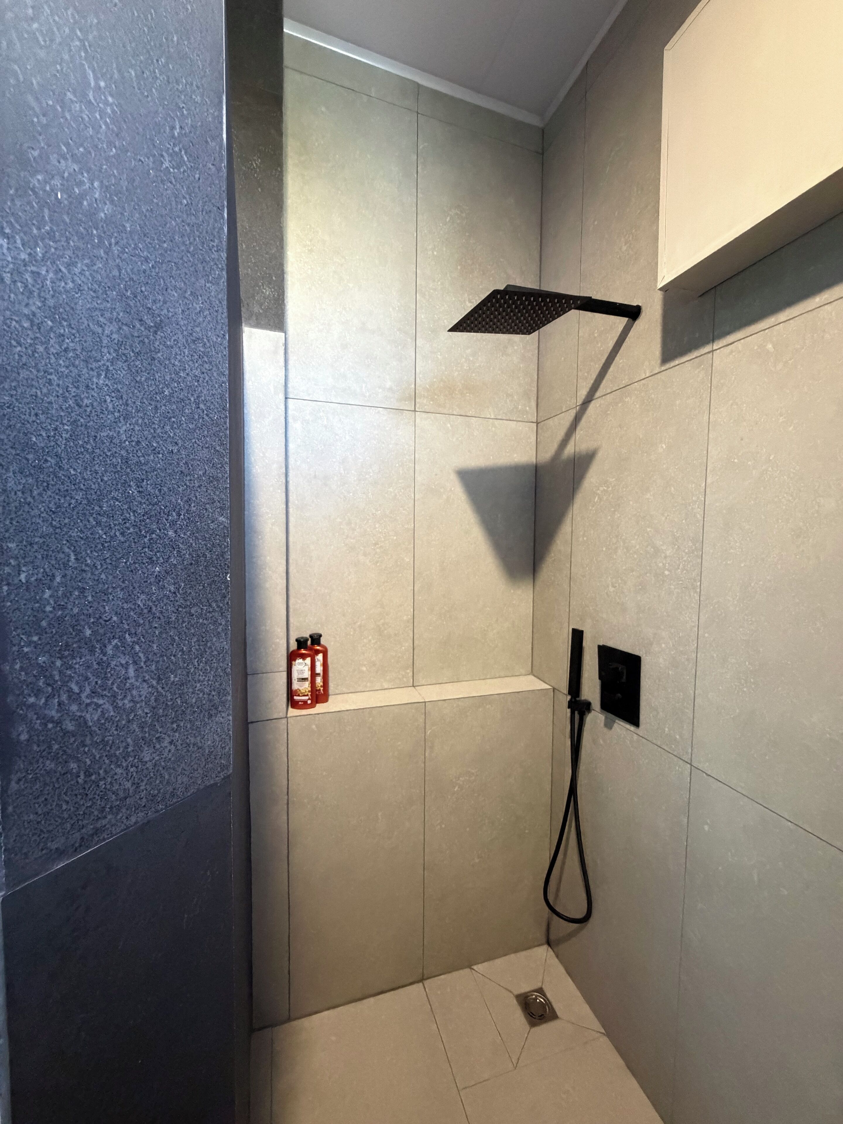 A shower area with a handheld showerhead, a small shelf with bottles, and a power outlet on the wall.