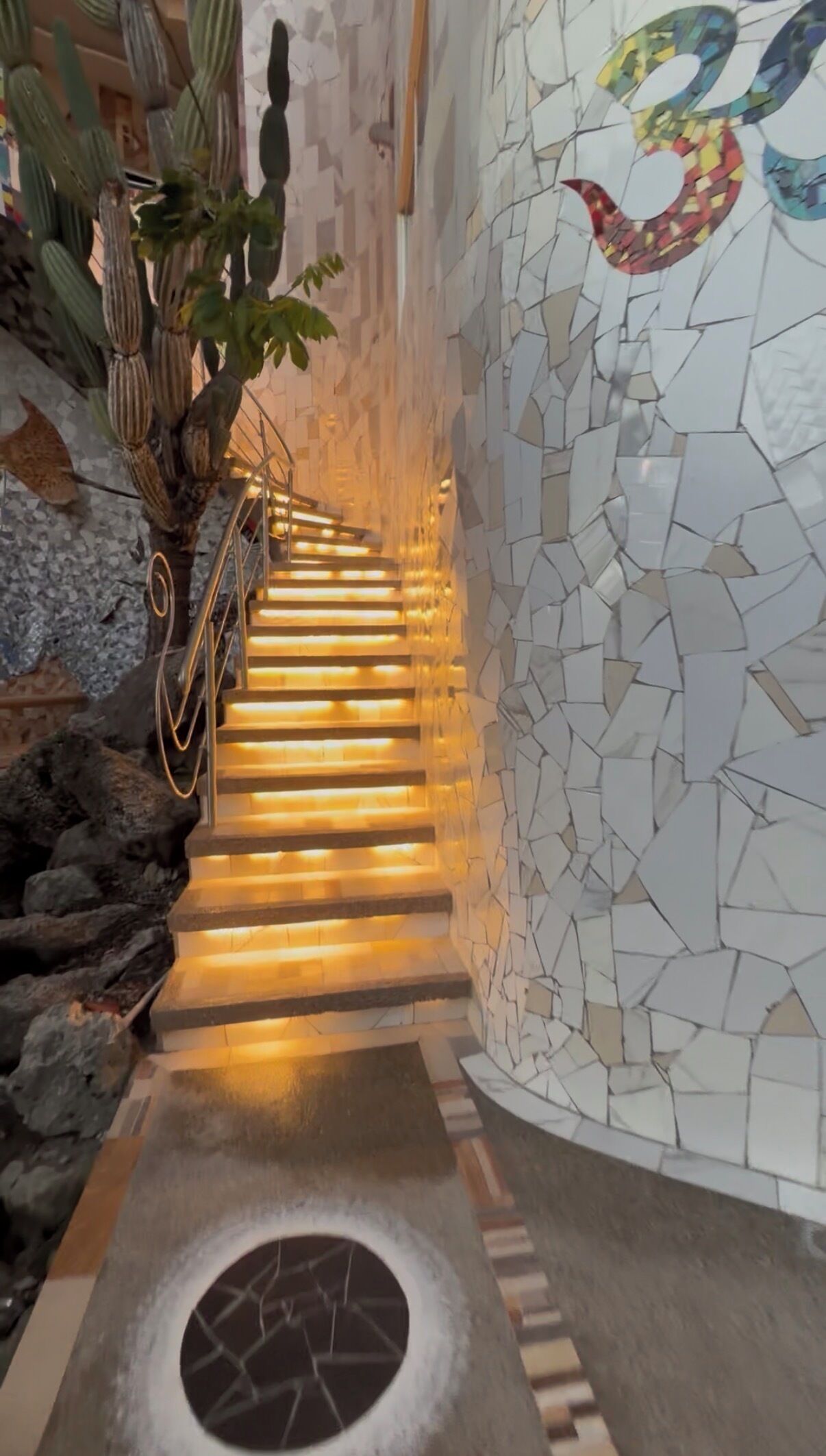 A staircase with illuminated steps leading upwards, surrounded by a mosaic wall and a cactus plant.