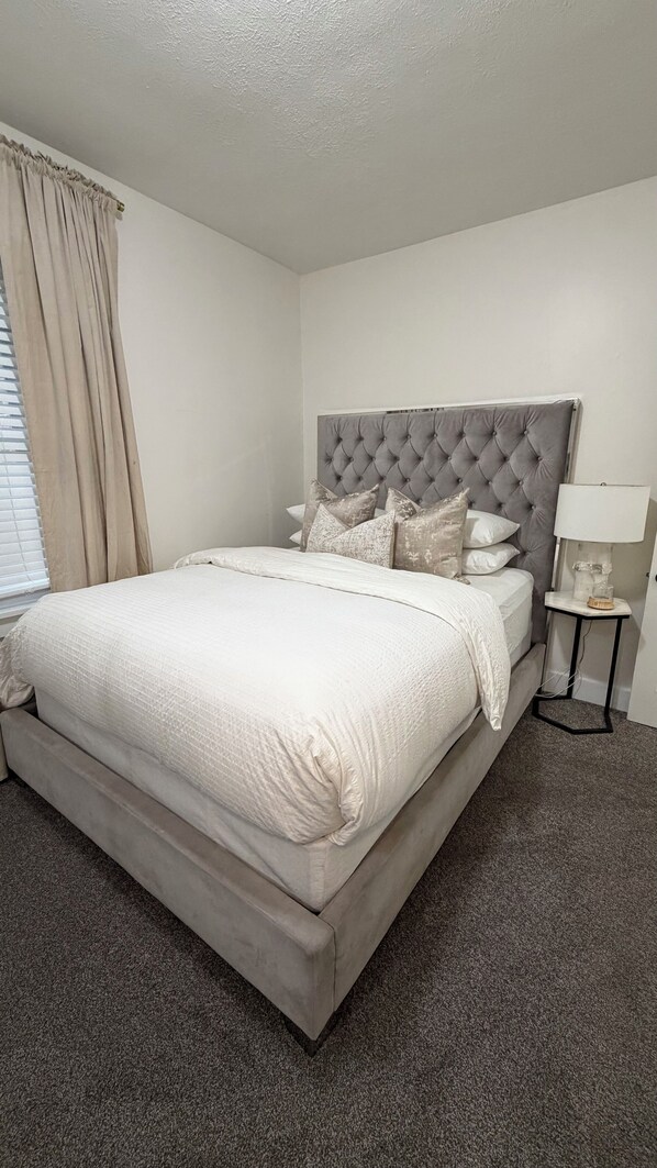 A neatly made bed with a tufted headboard, a bedside table with a lamp, and a window with curtains.