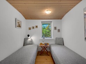 A small room with two single beds, a window with a view of greenery, a table with a vase, and a wall clock.