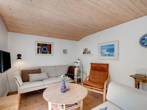 A cozy living room with a wooden ceiling, a white sofa, a brown leather chair, a round wooden table, and a TV mounted on the wall.