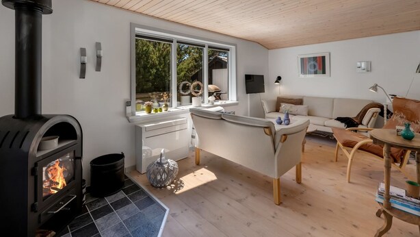 Ett modernt vardagsrum med en vedkamin, ett stort fönster, en vit soffa och en matplats.