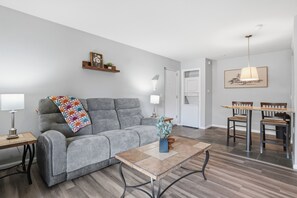 A modern living room with a grey sofa, a coffee table, a dining area with a table and chairs, and a kitchen visible through an open door. - Fully furnished, 1-bedroom condo in the heart of Tempe. (Tempe)