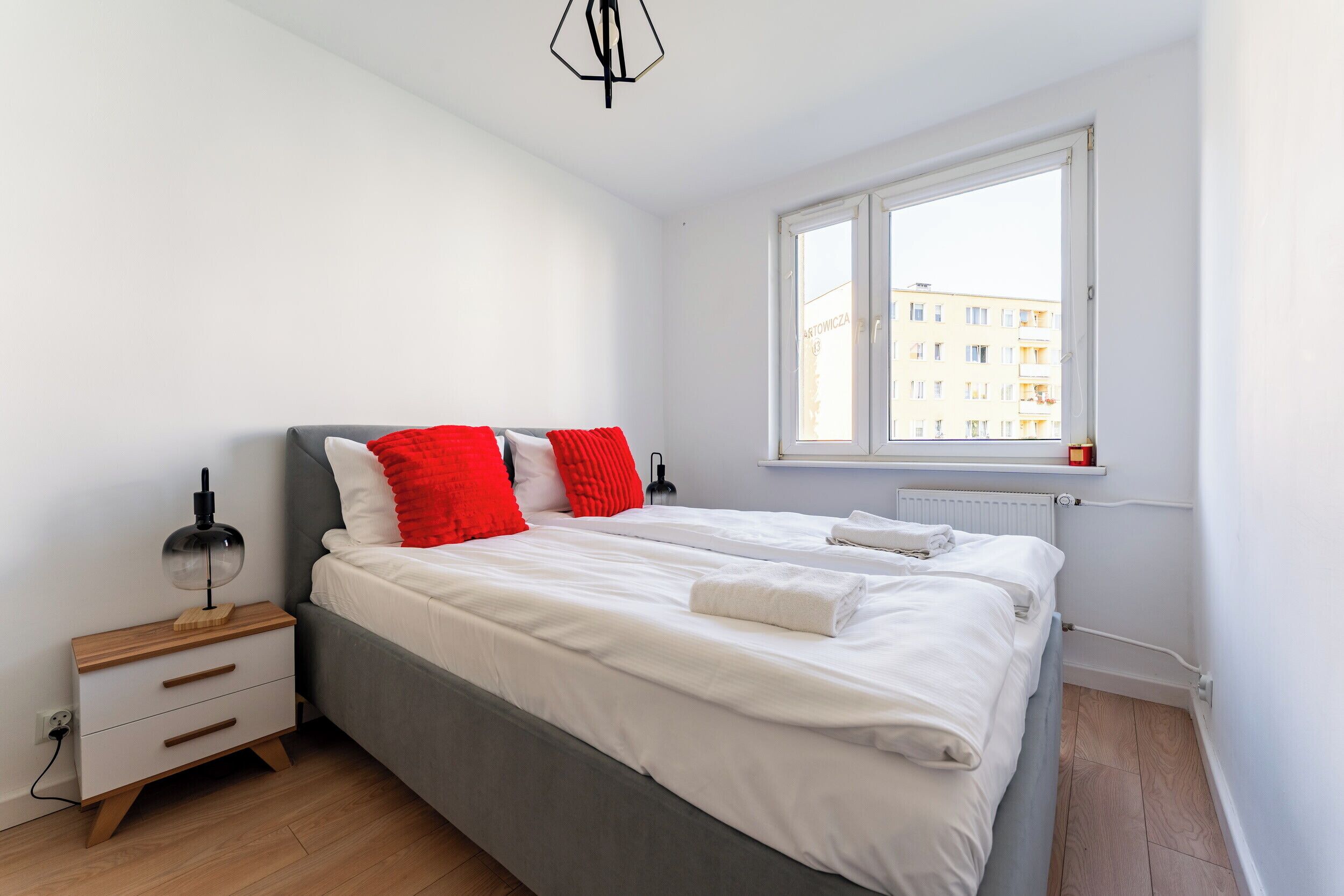 Ein modernes Schlafzimmer mit grauem Bett, weißer Bettwäsche und roten Kissen. Ein Nachttisch mit Lampe, ein Fenster mit Blick auf Gebäude und eine Heizung sind ebenfalls vorhanden.