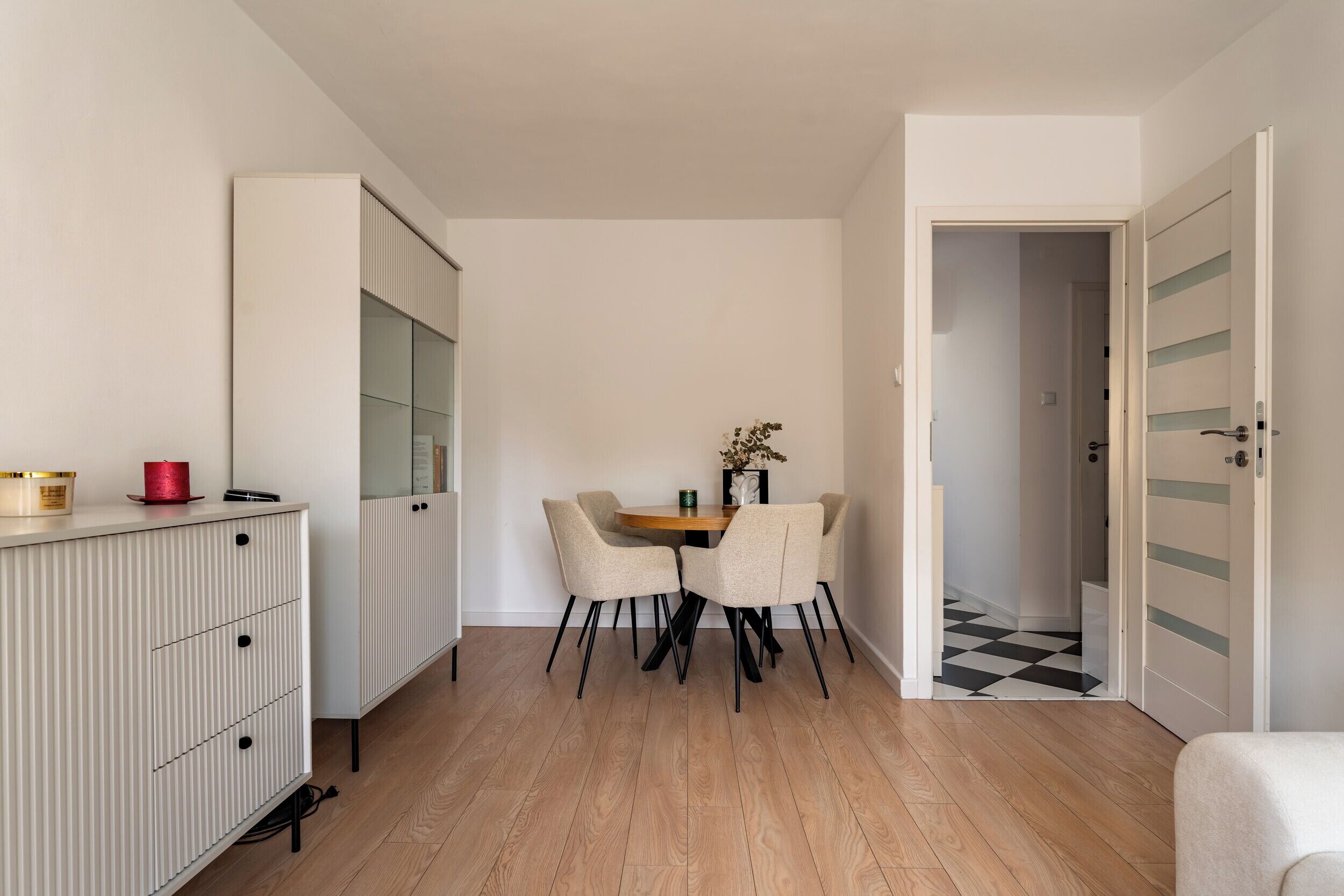 Ein moderner, minimalistischer Raum mit Holzboden, einem Esstisch mit Stühlen, einem weißen Schrank mit Glastüren und einer Tür zu einem weiteren Zimmer.