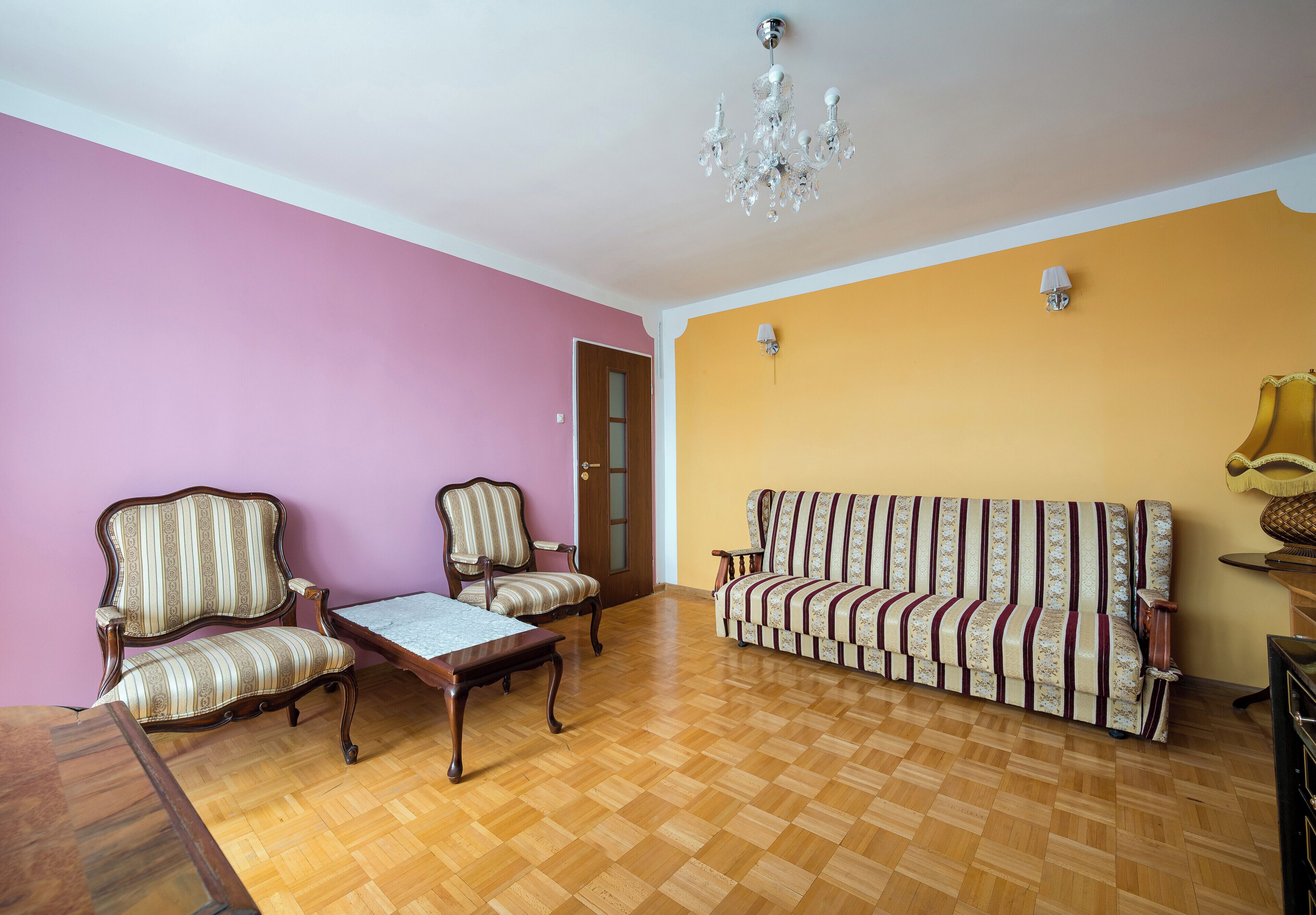 A room with a wooden parquet floor, two upholstered chairs, a sofa, a table, a chandelier, and a door.