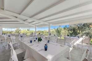 Un área de comedor al aire libre con mesas y sillas blancas, con árboles y un cielo despejado de fondo.