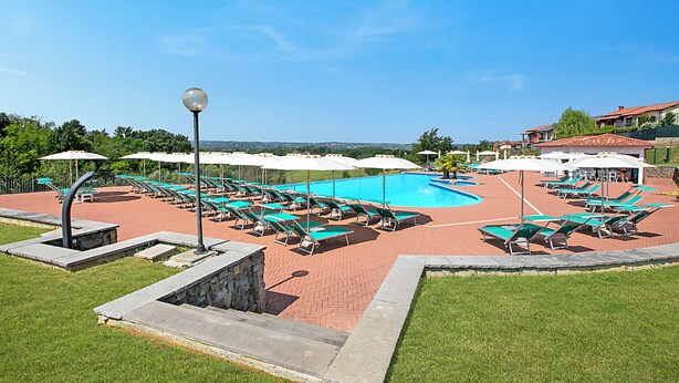 A pool area with multiple sun loungers and umbrellas, surrounded by a brick walkway and greenery.