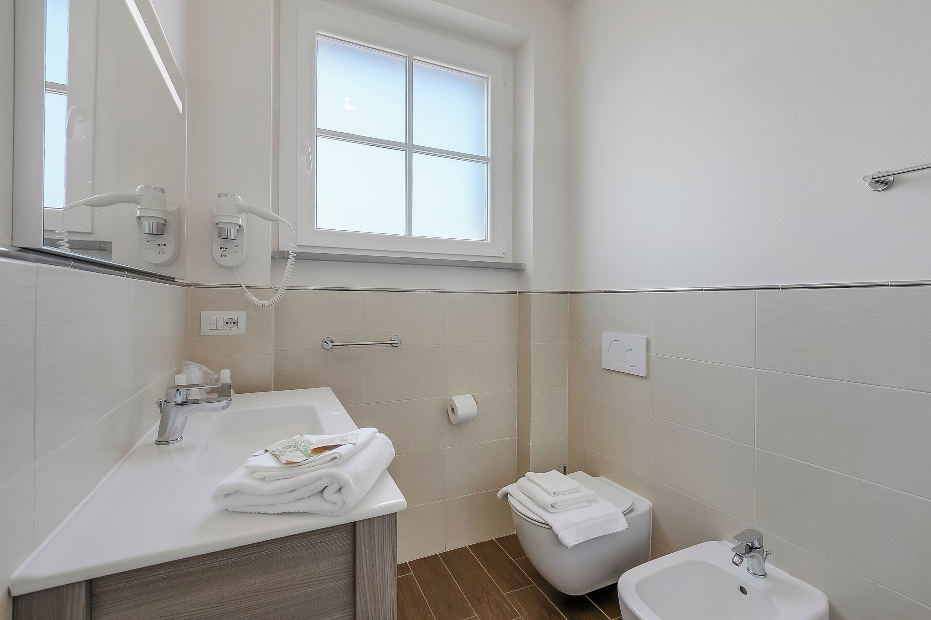 Een badkamer met een wastafel, spiegel, handdoekhouder, toilet, douchetoilet en een raam.